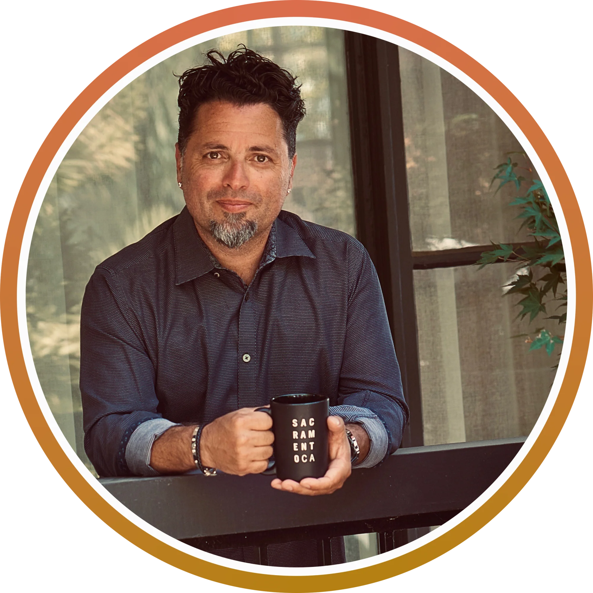 A man with dark curly hair, a goatee, and earrings holding a black mug with white letters that spell out 'SACRAMENTO' in a room with large windows and green plants.