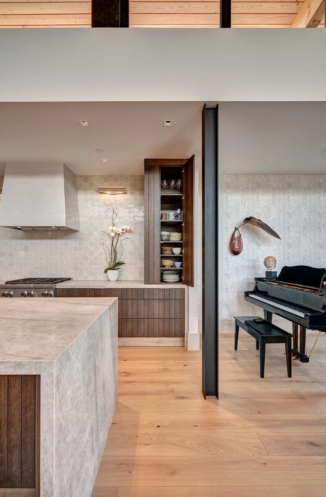 Interior of a modern home featuring a kitchen with wood accents, an orchid on the countertop, and a living area with a black grand piano, a decorative hanging vase, and a bust sculpture against light-colored walls.