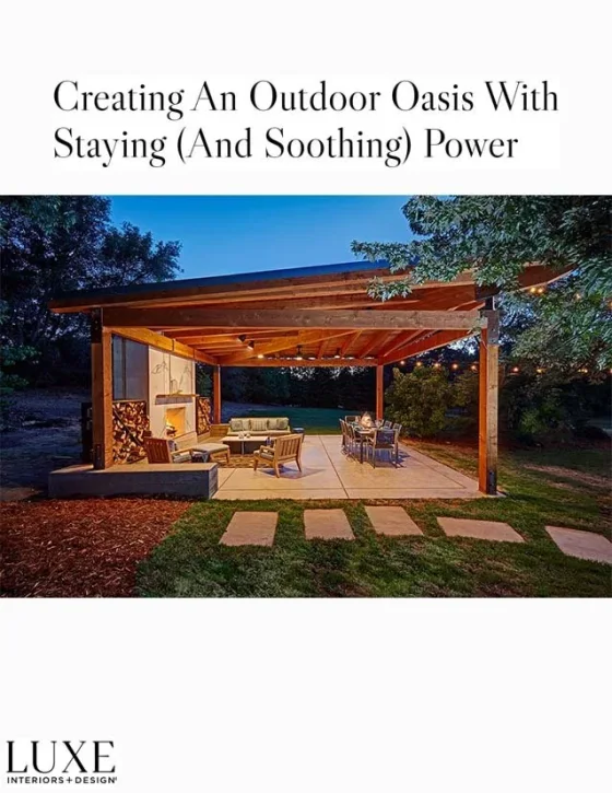 An outdoor covered patio with seating and dining furniture, surrounded by a lawn and trees at dusk.