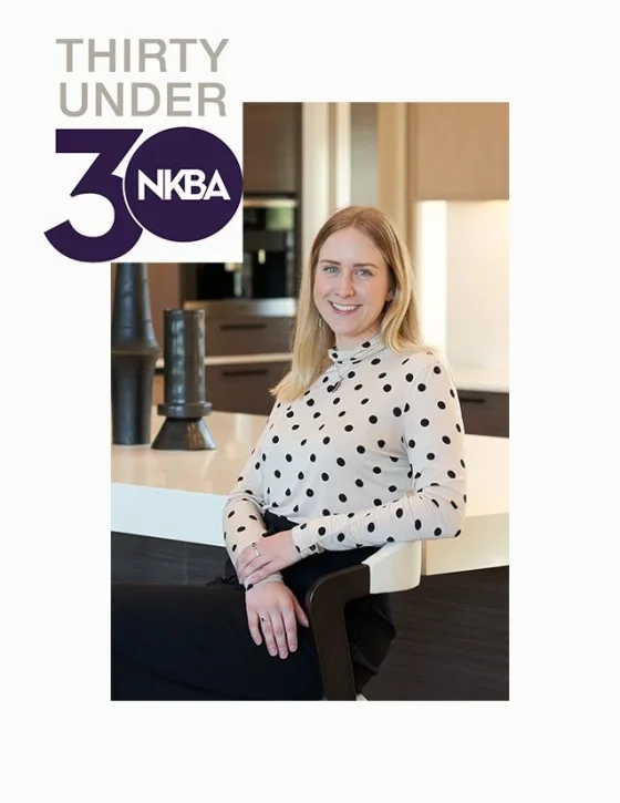 A woman with blonde hair wearing a white blouse with black polka dots, smiling, sitting in a modern kitchen with dark wood cabinets and decorative vases, celebrating NKBA's 30 Under 30.