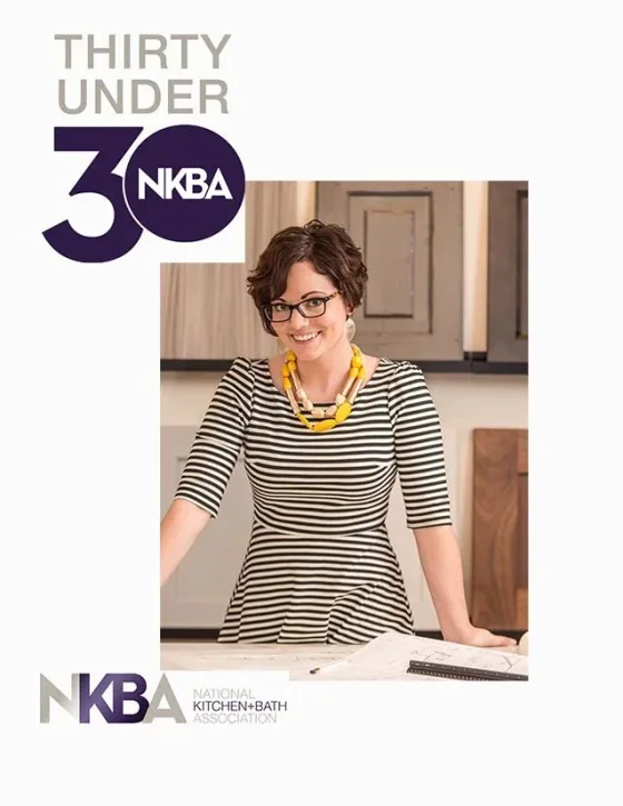 A woman wearing glasses and a striped dress with a yellow necklace, smiling and standing in a kitchen with cabinets, posing for a photo for the National Kitchen and Bath Association's 30 Under 30.